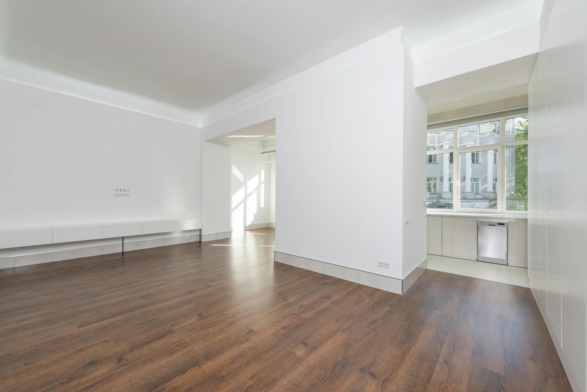 A contemporary empty apartment with white walls and wooden flooring, featuring large windows.